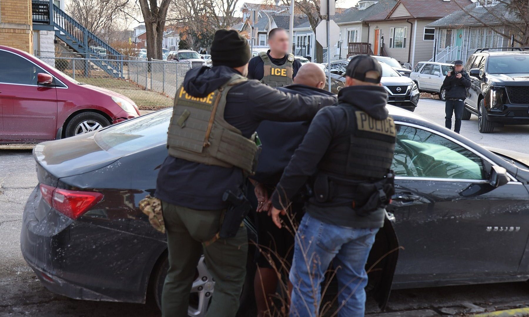 Uniformed ICE and police officers hold a man beside a dark car on a residential street, with faces blurred