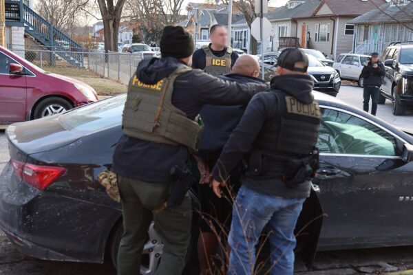 Uniformed ICE and police officers hold a man beside a dark car on a residential street, with faces blurred