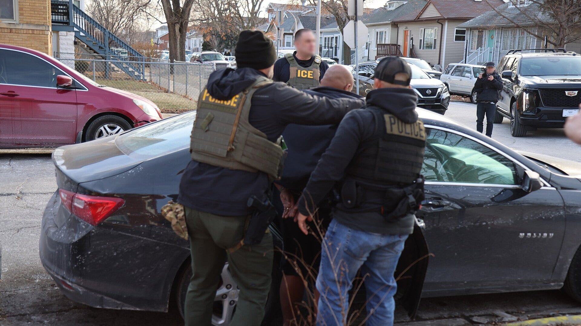 Uniformed ICE and police officers hold a man beside a dark car on a residential street, with faces blurred