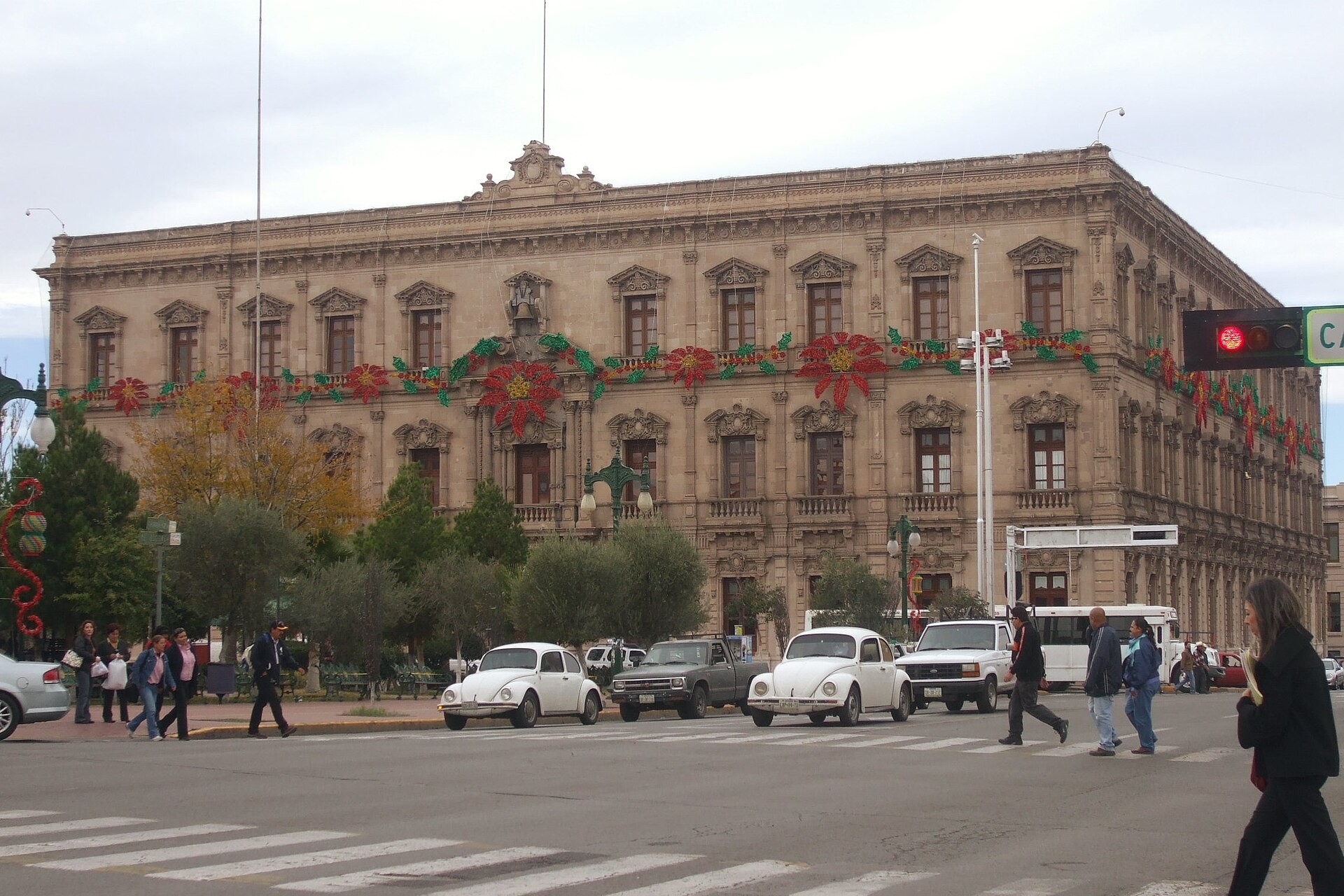 The Government Palace in Chihuahua City, a sandstone building with holiday decorations on its facade, with cars and pedestrians crossing the street in front