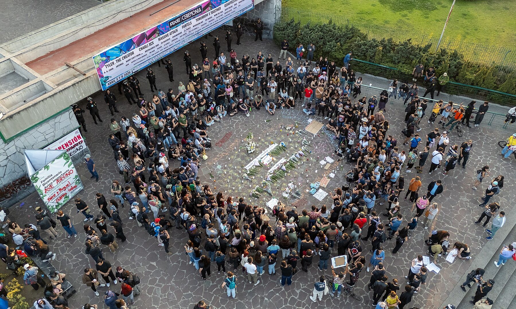 Aerial view of a large crowd gathered around candles and flowers at a vigil on a plaza in Parque Bicentenario, Mexico City