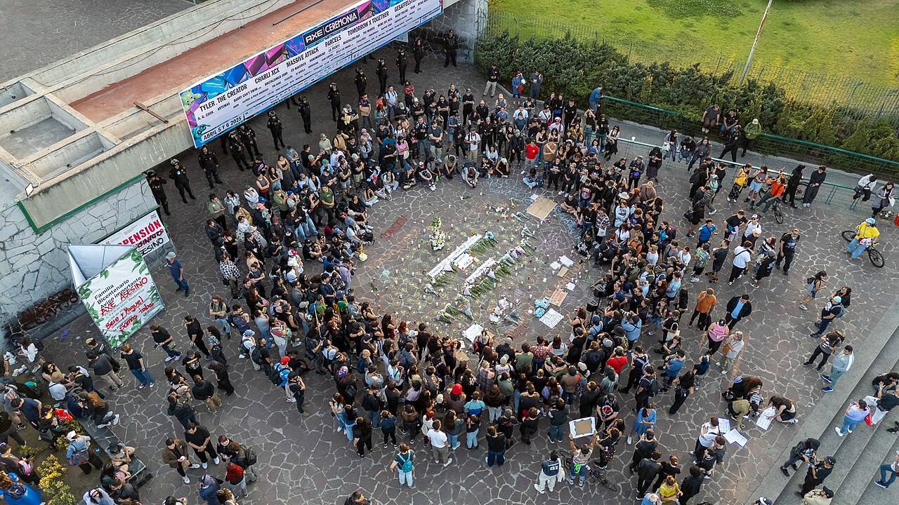 Aerial view of journalists and relatives gathered around flowers and candles in Parque Bicentenario, Mexico City, beneath an Axe Ceremonia festival banner