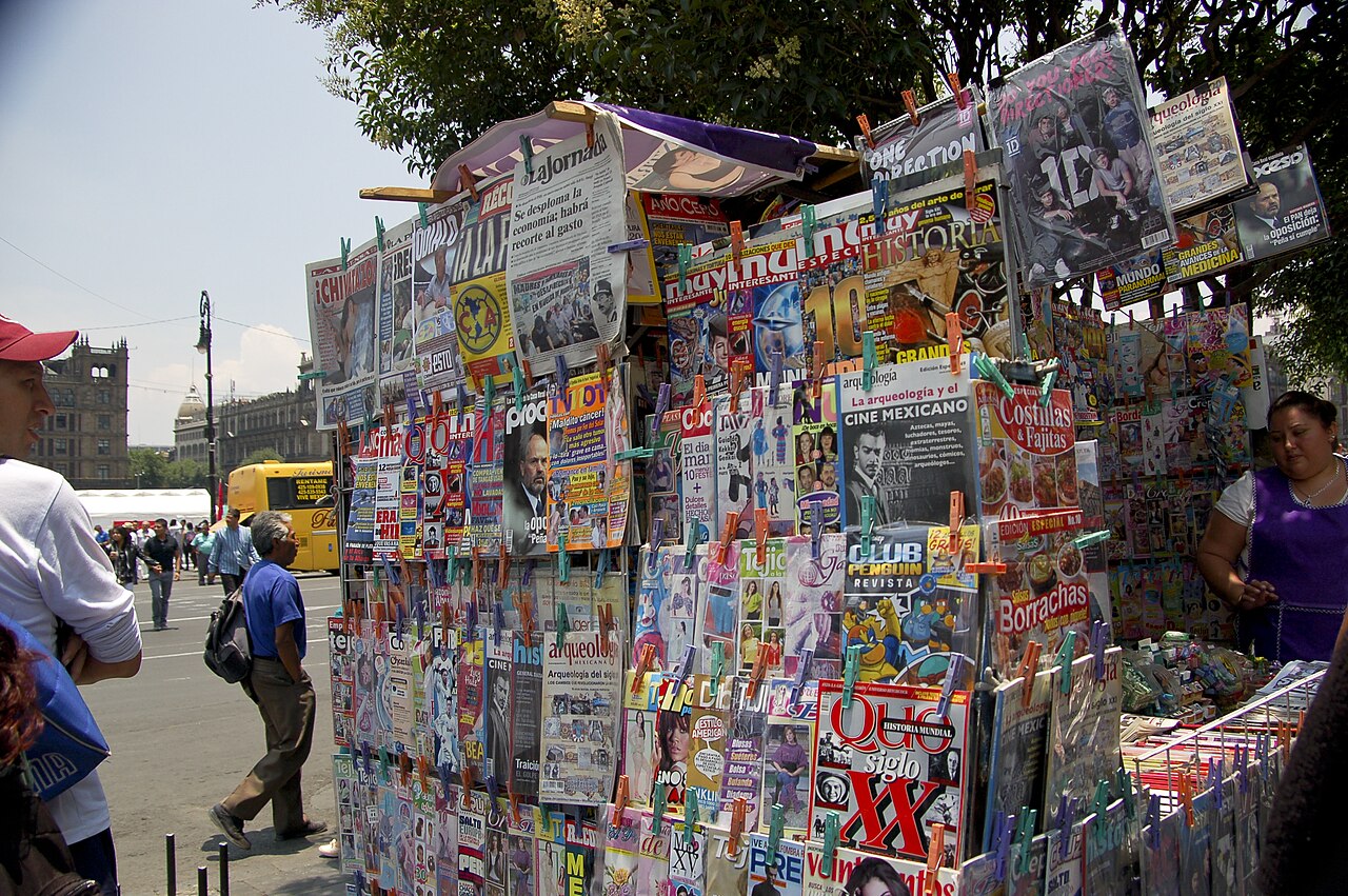 A street newsstand in Mexico City hung with newspapers and magazines, including La Jornada, with a vendor beside it