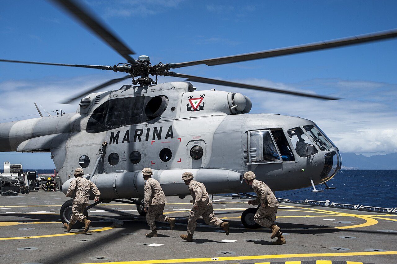 Grey Mexican Navy Mi-17 helicopter marked MARINA with rotors turning on a ship deck, with US Marines running past under a blue sky