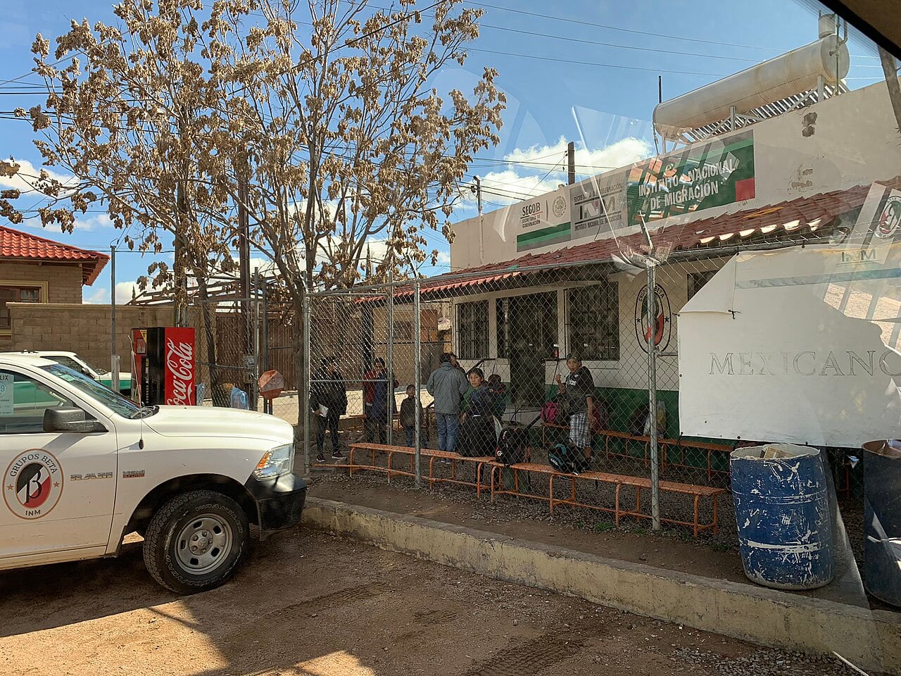 People wait behind a chain-link fence outside an office of Mexico's National Migration Institute, with an INM Grupos Beta pickup truck parked in front