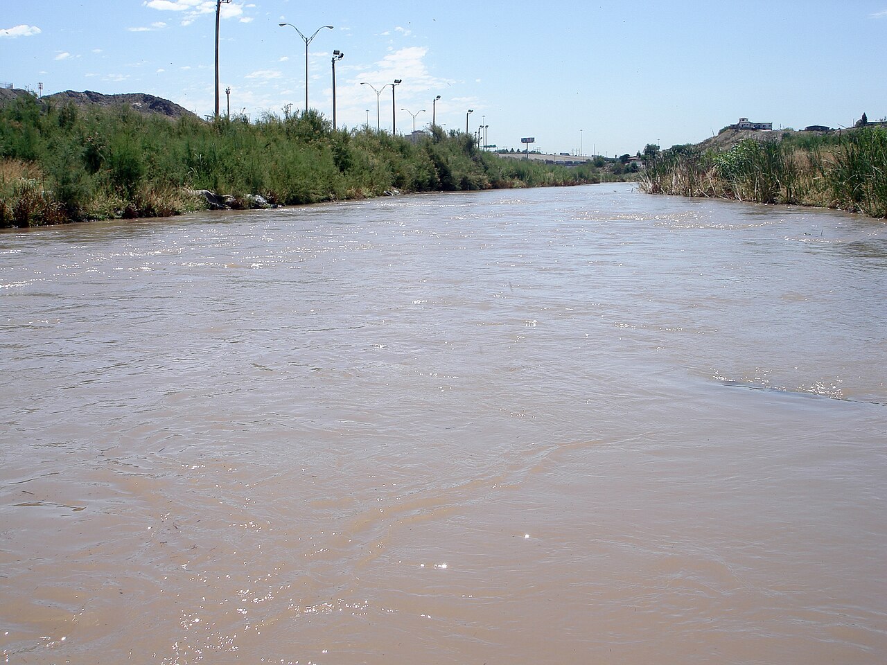 The brown waters of the Rio Grande flow between reed-covered banks, with street lights and a highway along one side