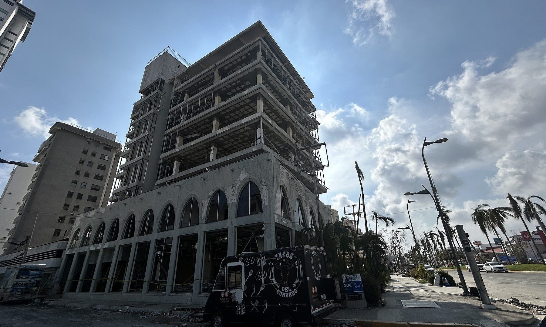 A high-rise building in Acapulco stripped of its windows and facade by Hurricane Otis, with damaged palm trees along the avenue