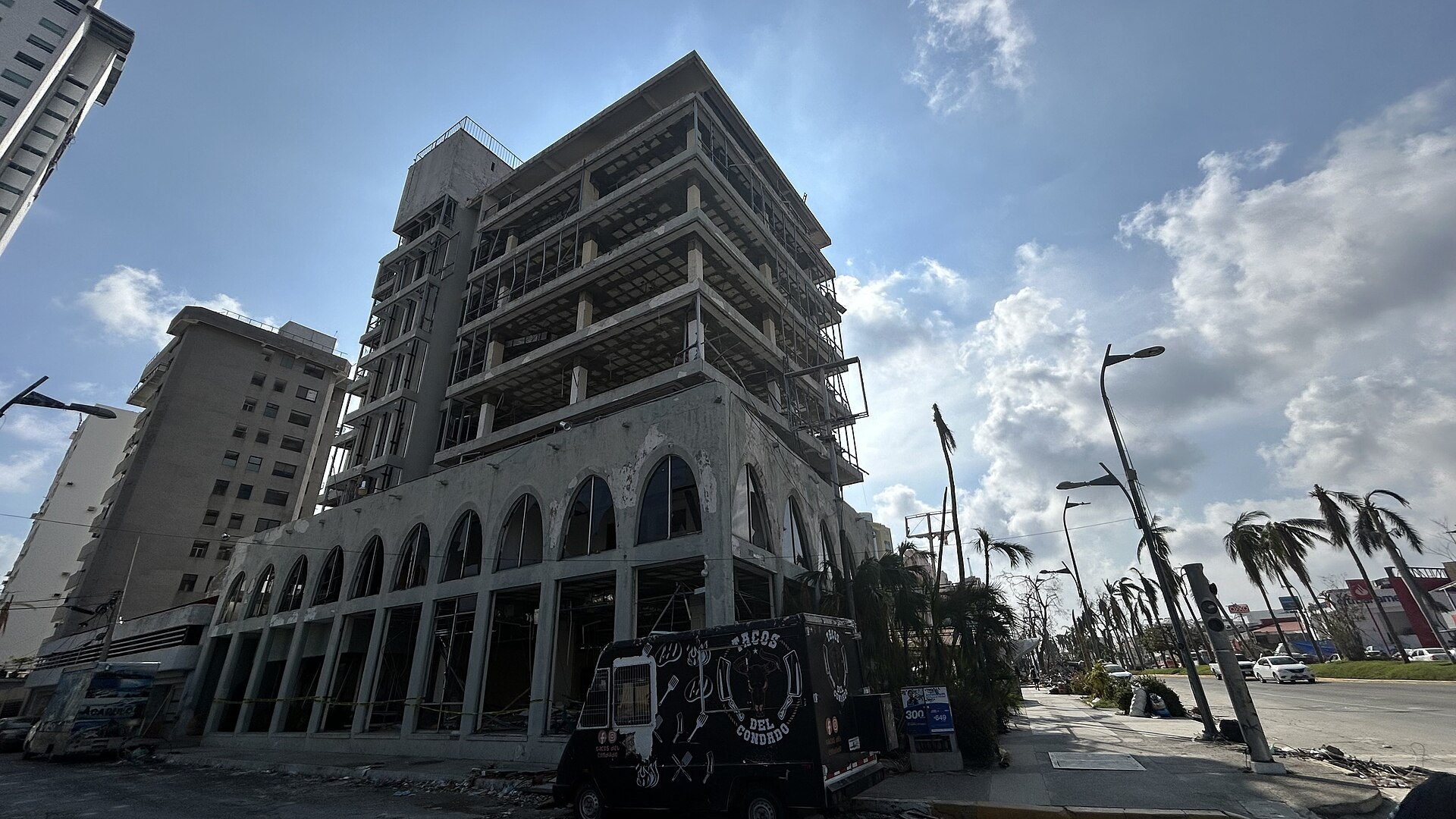 A high-rise building in Acapulco stripped of its windows and facade by Hurricane Otis, with damaged palm trees along the avenue