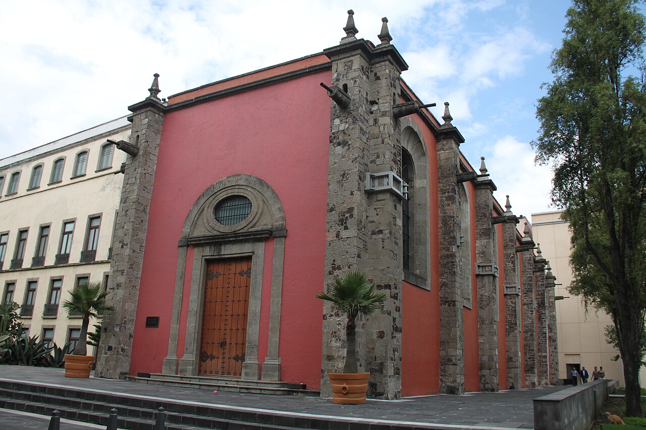 Red-walled stone building with a carved wooden door and potted palms, part of the National Palace complex in Mexico City