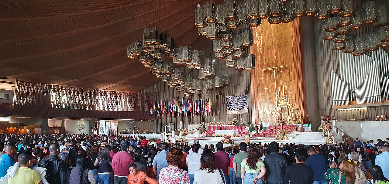 Crowd of worshippers inside the modern Basilica of Guadalupe in Mexico City, with flags and organ pipes near the altar