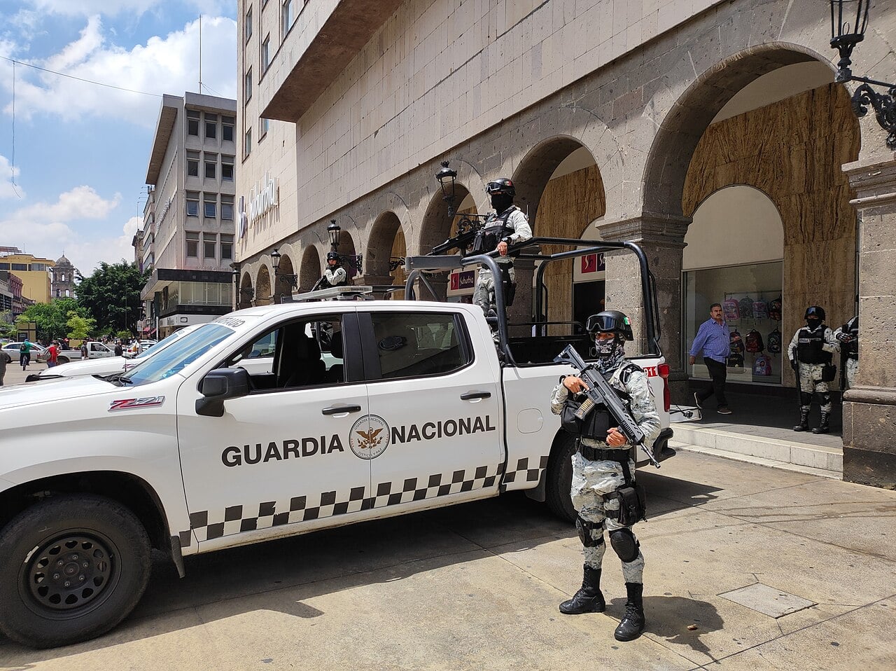 Masked National Guard members in camouflage, holding rifles, stand beside a white Guardia Nacional pickup truck on a city street