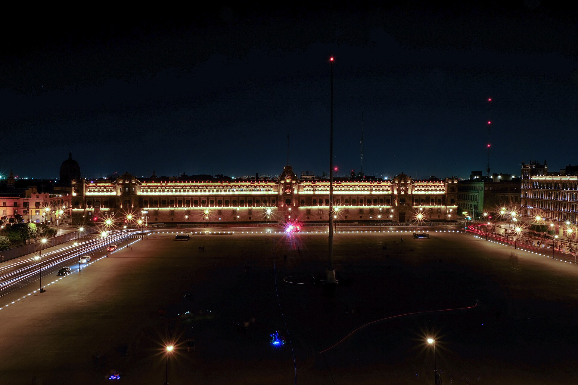 The National Palace in Mexico City, lit at night beyond the empty Zócalo square, with a tall flagpole at the centre