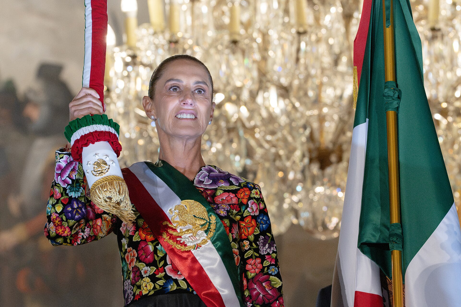 Mexican President Claudia Sheinbaum, in an embroidered floral blouse and the presidential sash, holds a ribbon-wrapped flag staff beside the Mexican flag at the Grito de Independencia ceremony