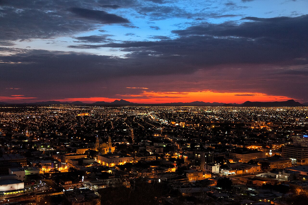 Hermosillo seen from above at dusk, with city lights spreading to distant mountains under a red sunset and dark clouds