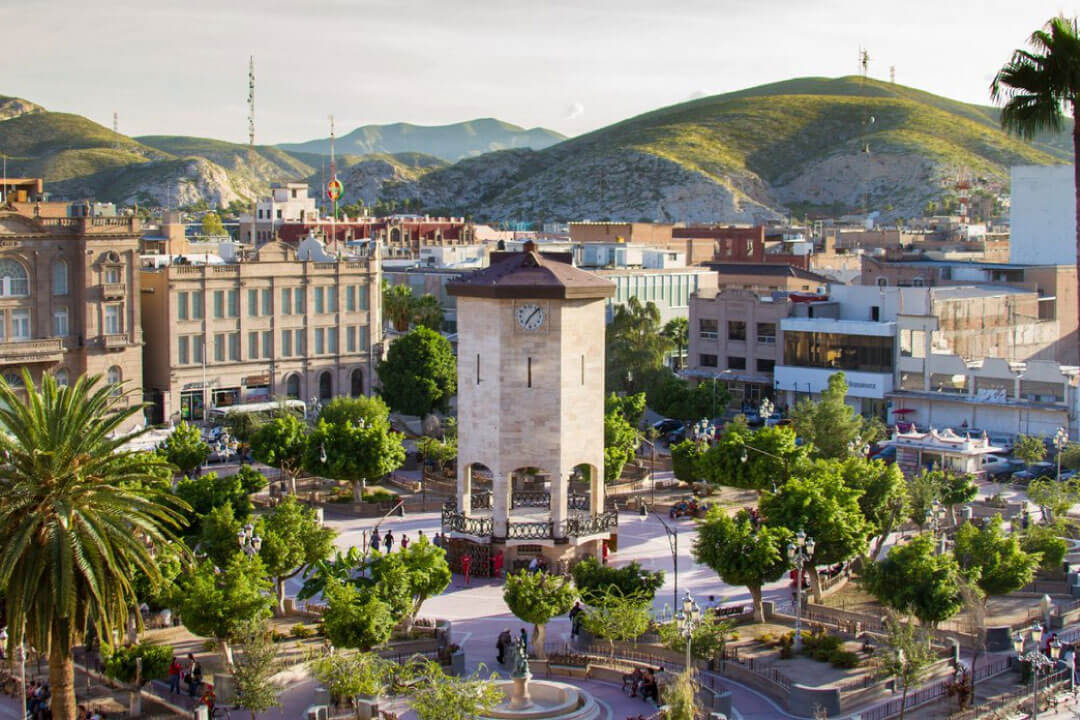Aerial view of a square with a stone clock tower, palm trees and paths, surrounded by city buildings and dry hills