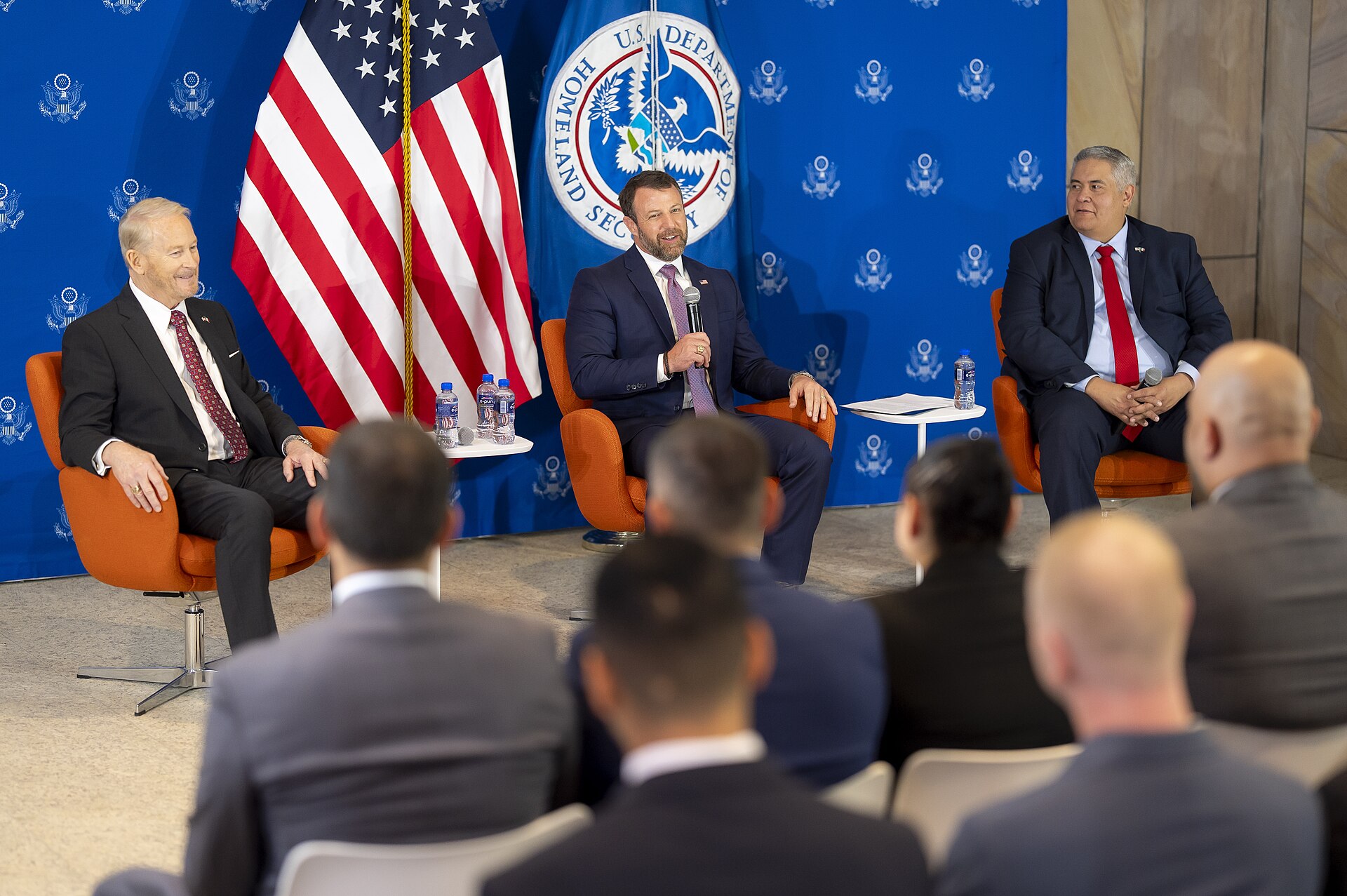 US Homeland Security Secretary Markwayne Mullin speaking into a microphone on stage at a town hall at the US Embassy in Mexico City, with two men seated beside him