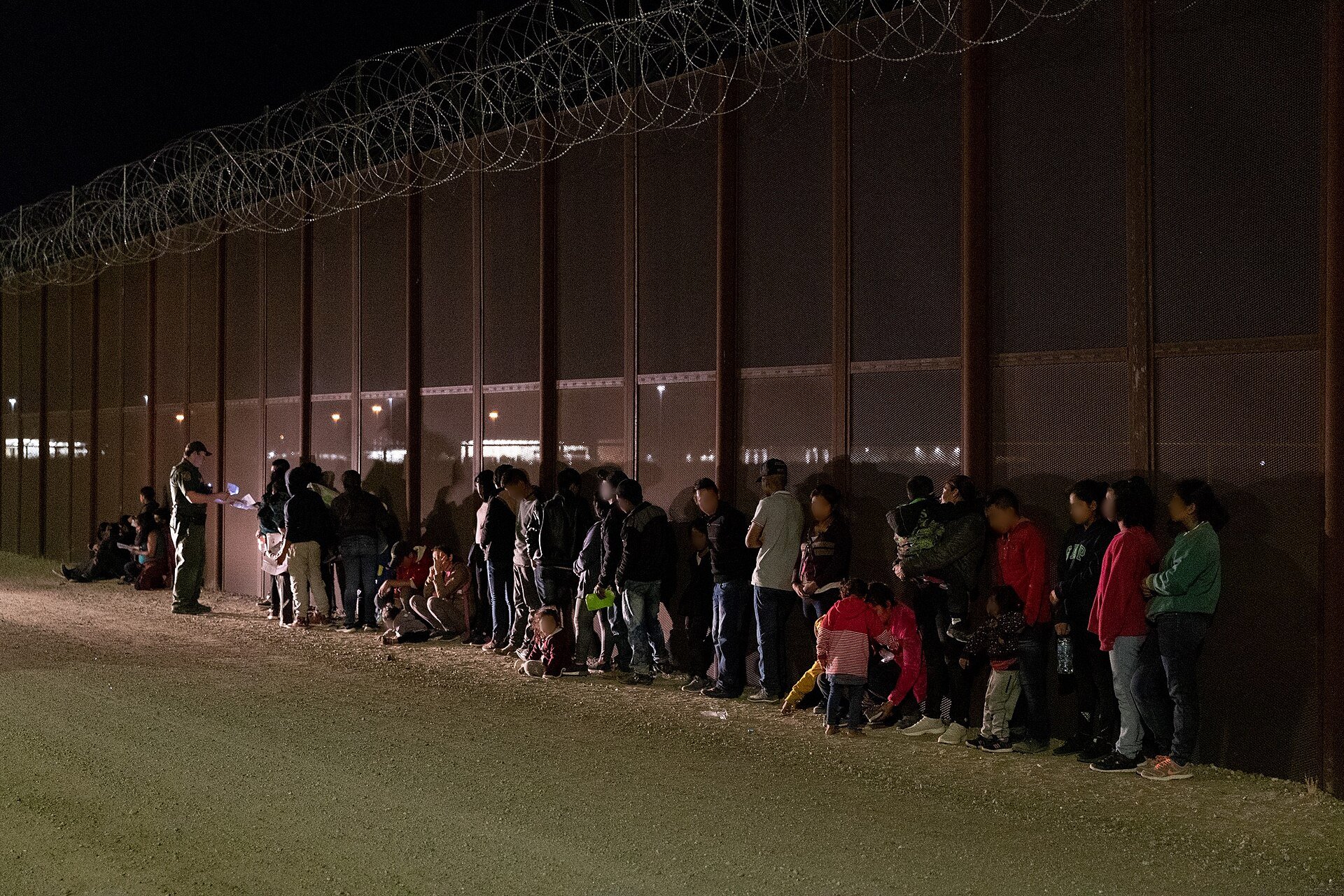 Migrants lined up beside the tall border wall at night while a Border Patrol agent checks papers near Yuma, Arizona