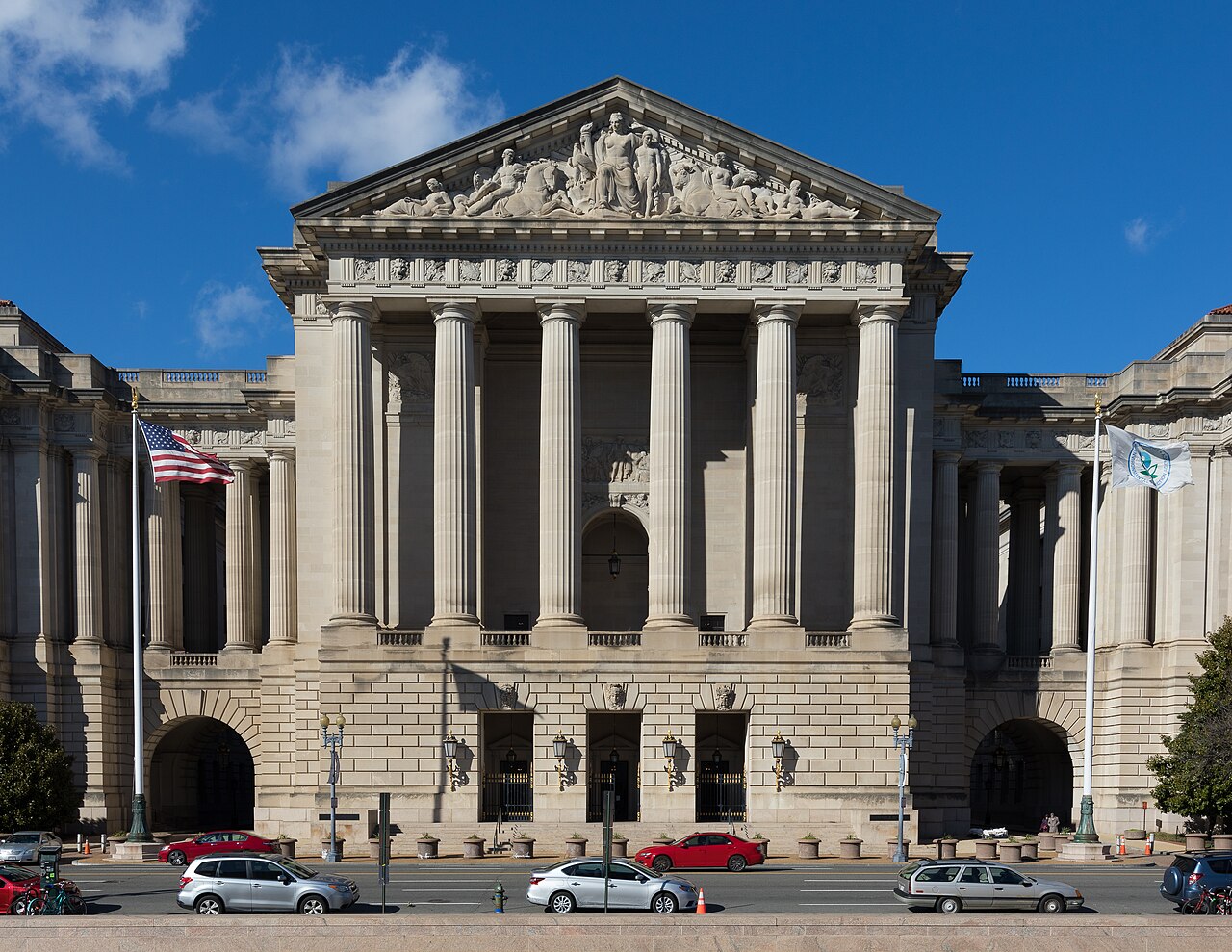 The columned neoclassical facade of the Andrew W. Mellon Auditorium in Washington under a blue sky, with a US flag flying in front