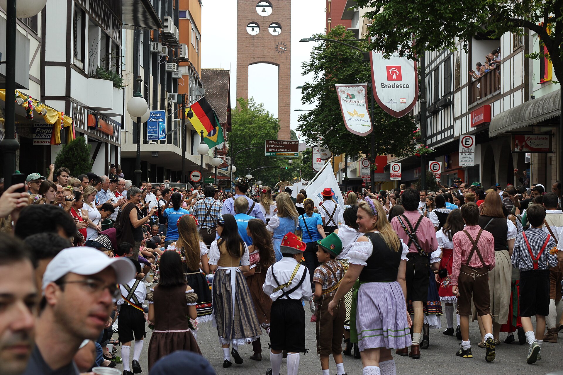 Crowd in traditional German-style dress fills a Blumenau street lined with half-timbered buildings, with a brick tower in the background