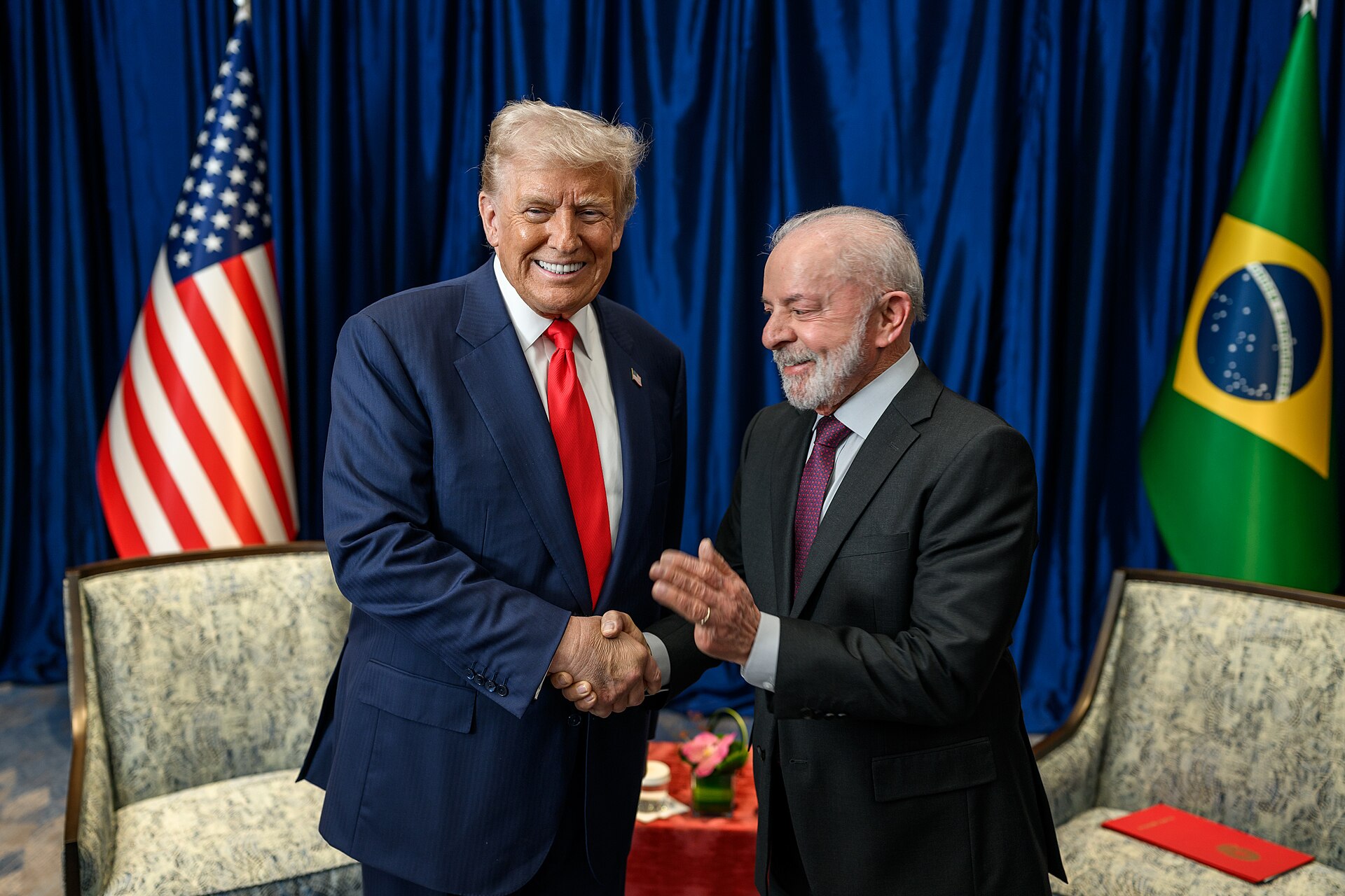 President Trump and President Lula shake hands and smile in front of a US flag and a Brazilian flag, with blue curtains behind them