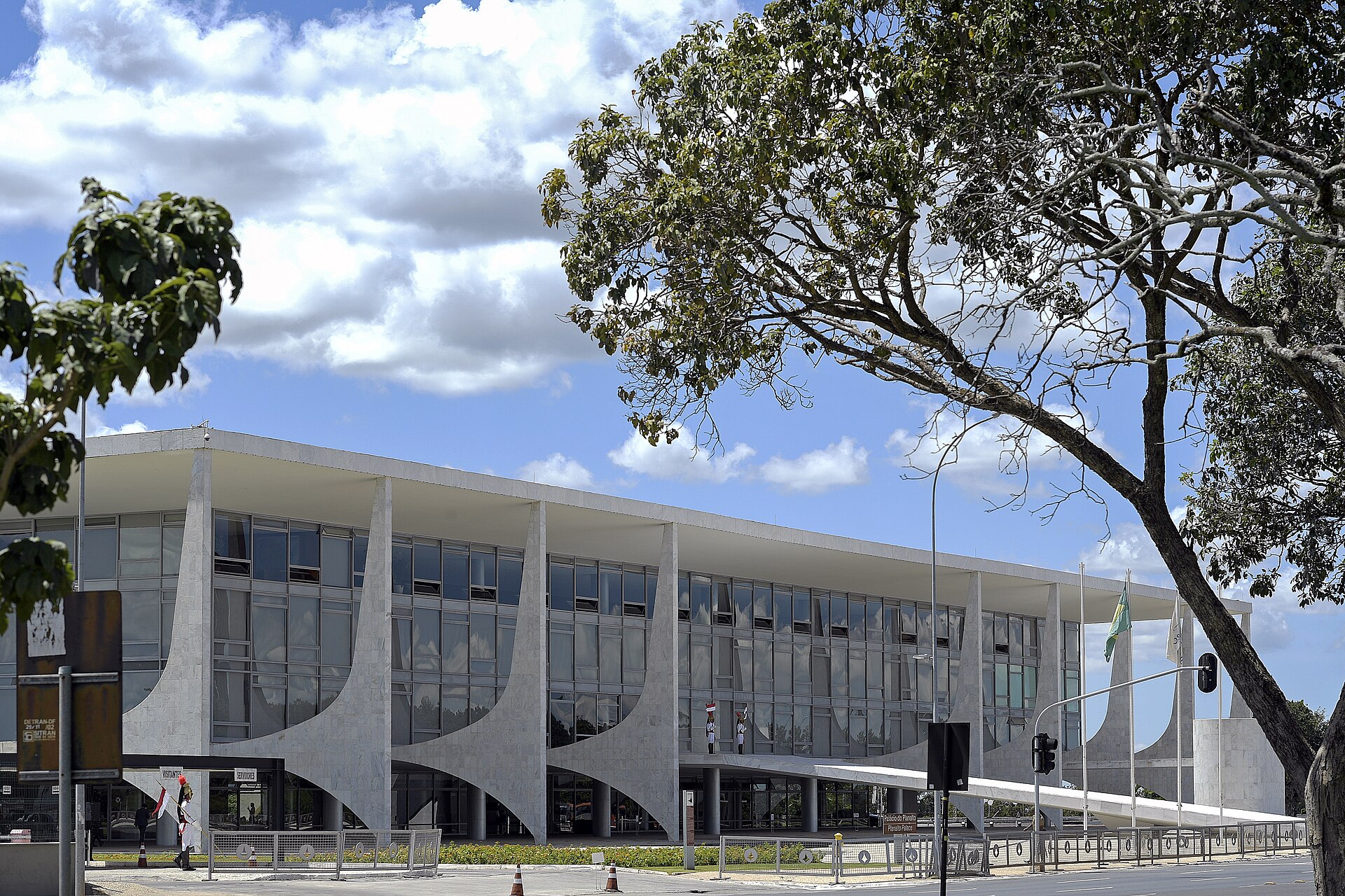 Modern white concrete facade of the Palácio do Planalto with tall columns, glass walls and a tree in the foreground under a blue sky