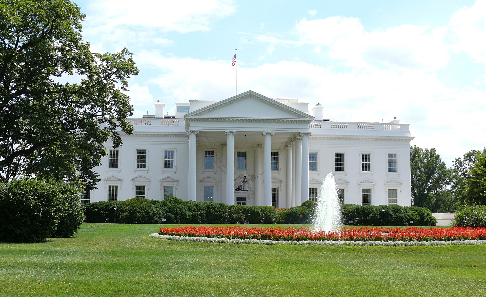 The north front of the White House with white columns and a flag, a fountain and red flowerbed on the lawn in front under a blue sky