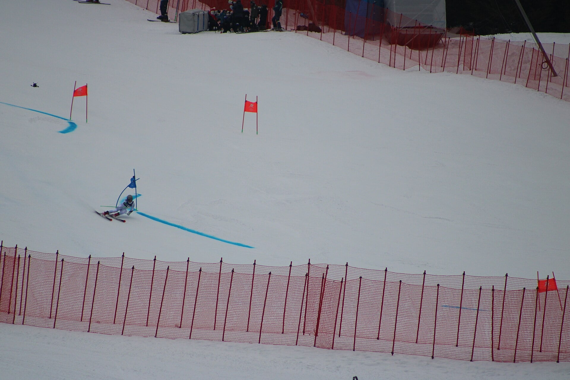 A skier in a white race suit carves through a blue-marked turn on a snow slope between red and blue gates, with orange safety netting in the foreground