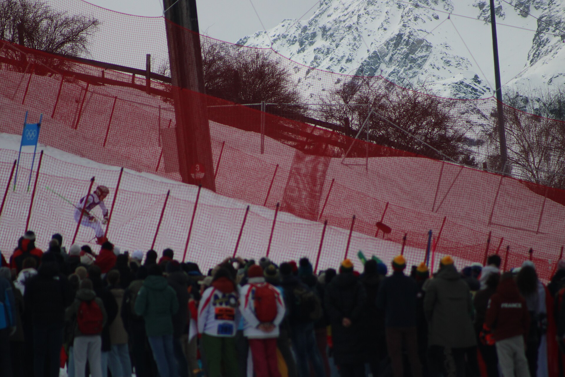 A skier in a red and white race suit passes red safety netting above a crowd of spectators, with snow-covered mountains behind