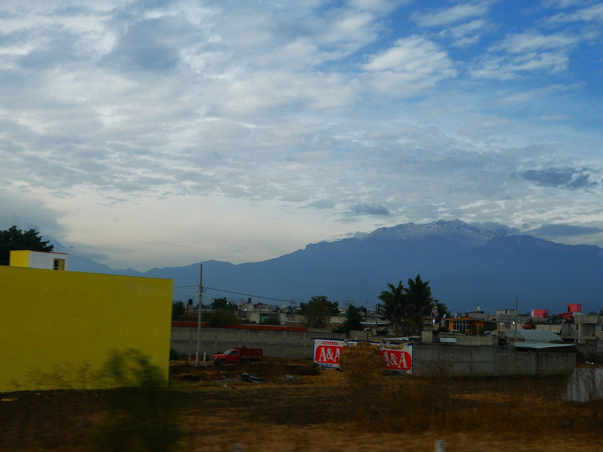 Snow-dusted Iztaccihuatl volcano rising behind low buildings and a roadside lot under a cloudy sky, seen from the Mexico City to Puebla highway