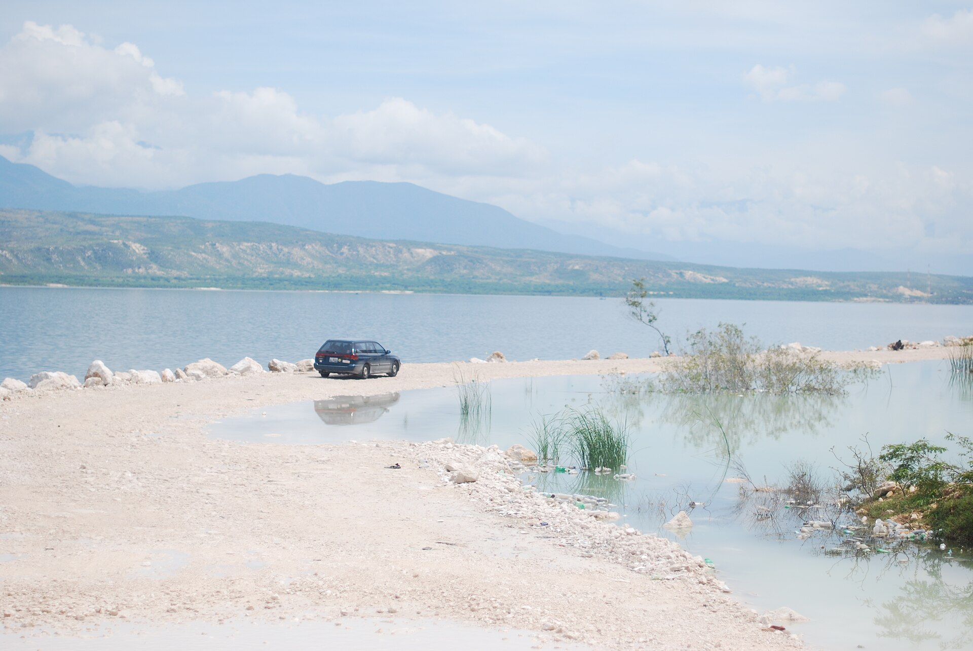 A car parked on a pale shoreline road beside a flooded lake with hills in the distance under a cloudy sky