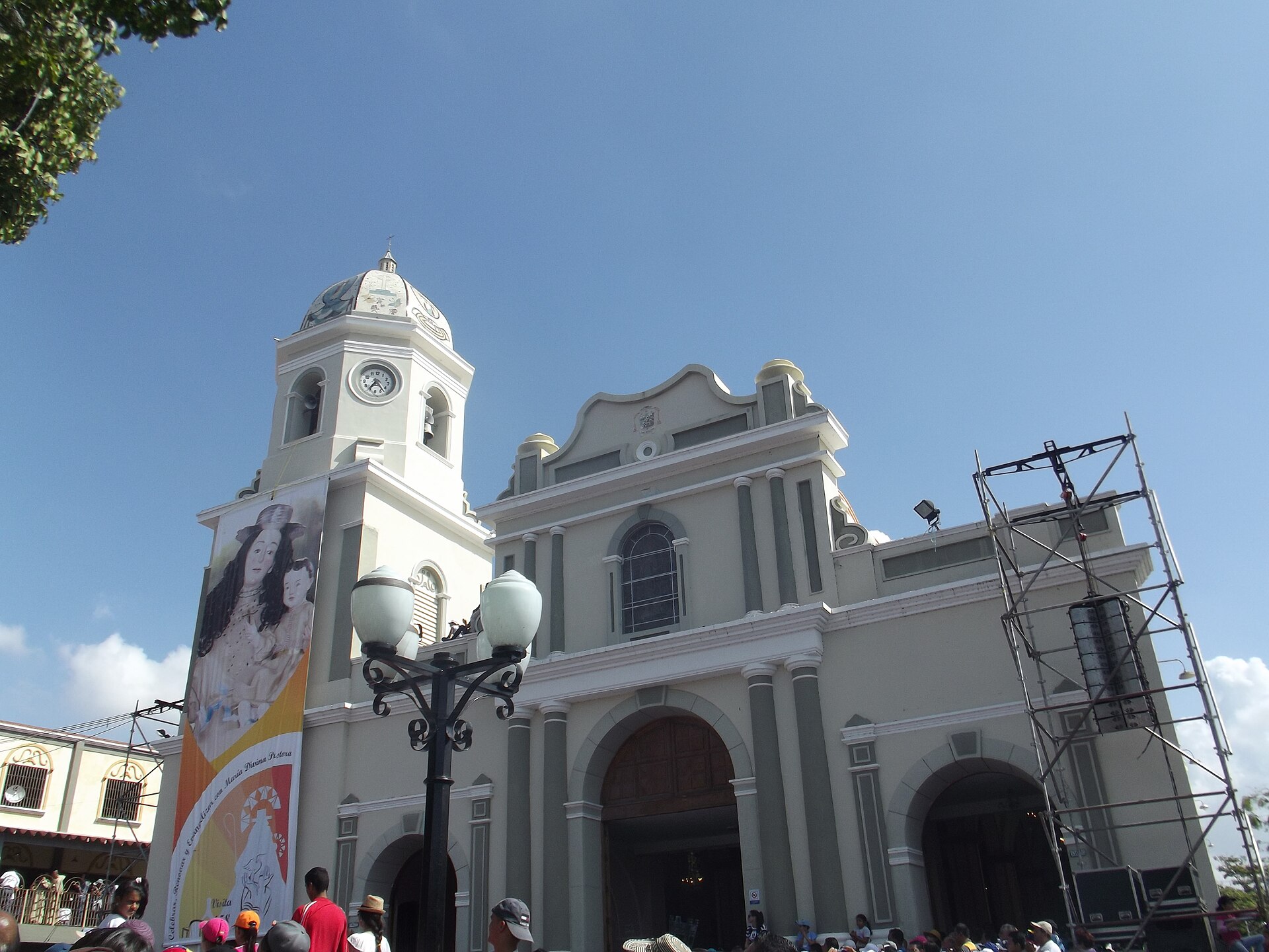 The cream and green facade of the Basilica of Santa Rosa in Barquisimeto under a blue sky, with a crowd gathered below and a stage scaffold at right