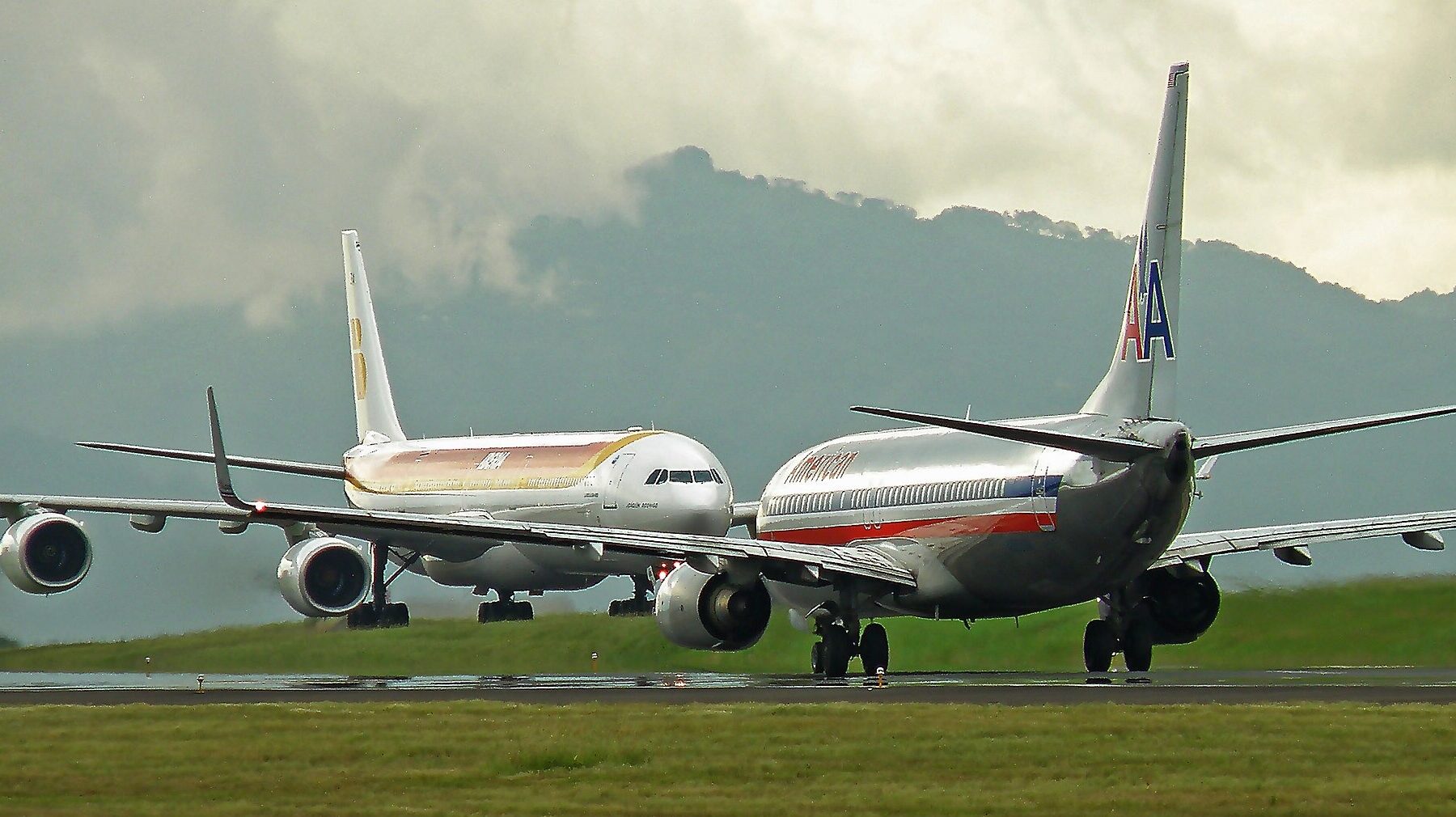 An Iberia Airbus and an American Airlines Boeing 737 on the runway at Juan Santamaria airport with green hills and cloud behind