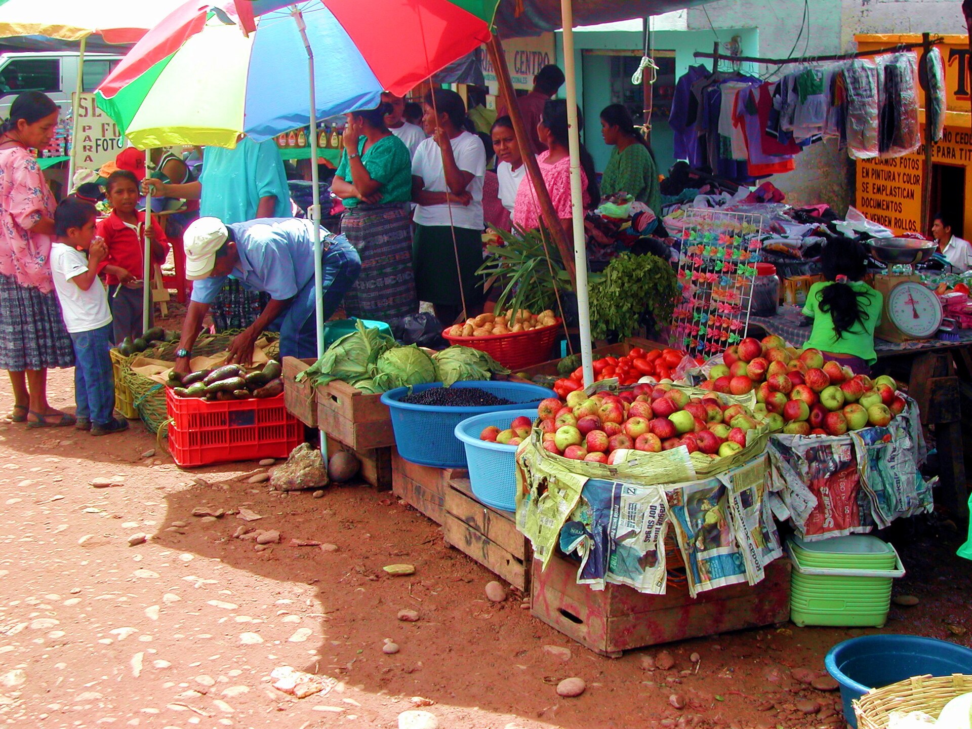 Open-air market stalls under a striped umbrella in Playa Grande with fruit, vegetables and shoppers, including women in traditional skirts