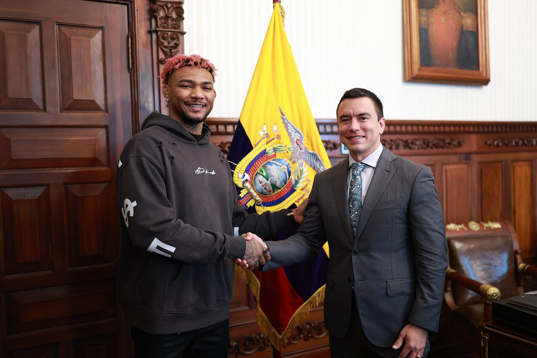 Ecuadorian UFC fighter Michael Morales, in a dark hooded sweatshirt, shakes hands with President Daniel Noboa in front of the Ecuadorian flag