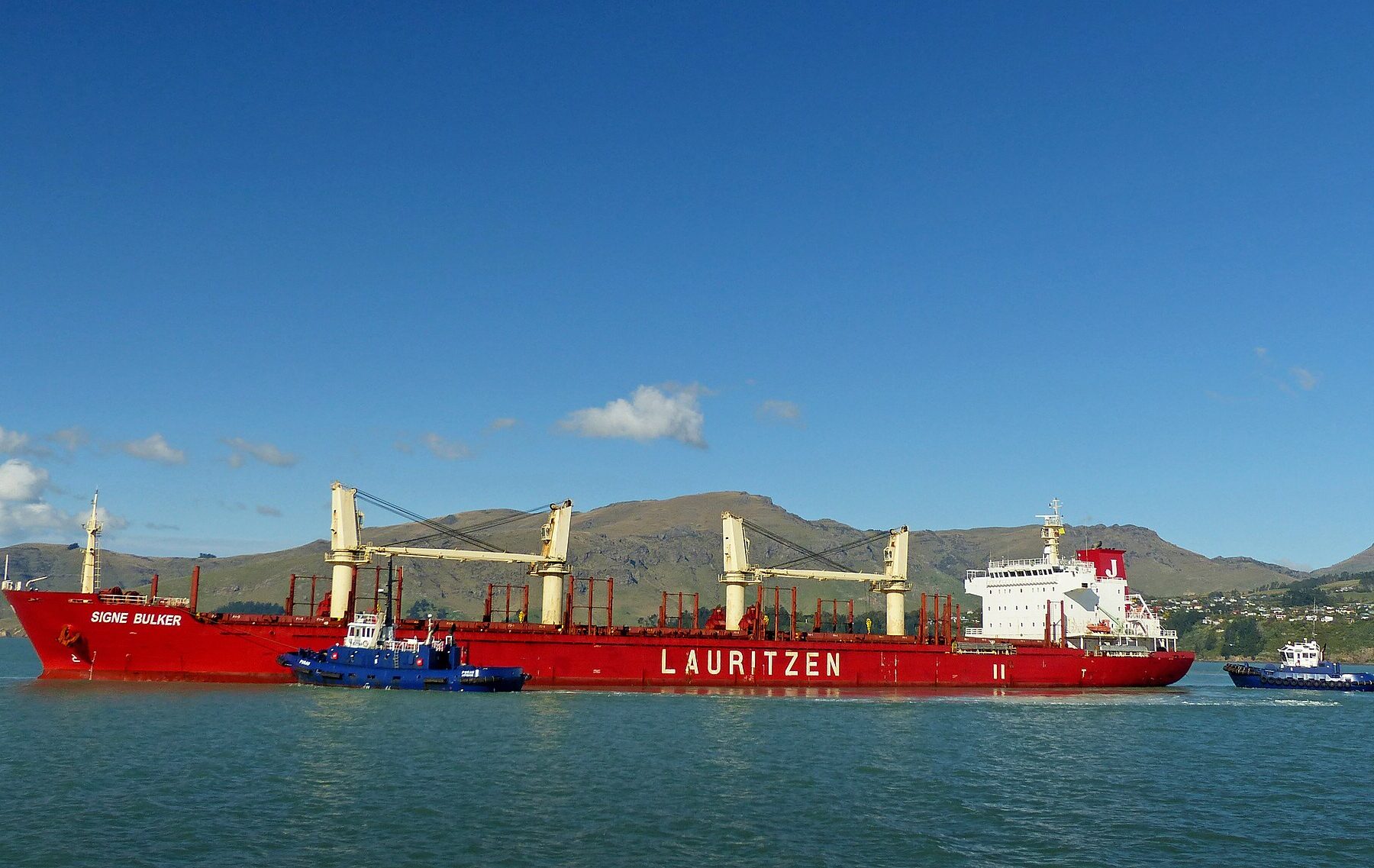 A red bulk carrier with yellow cranes and the name Signe Bulker, escorted by two tugboats, with hills behind