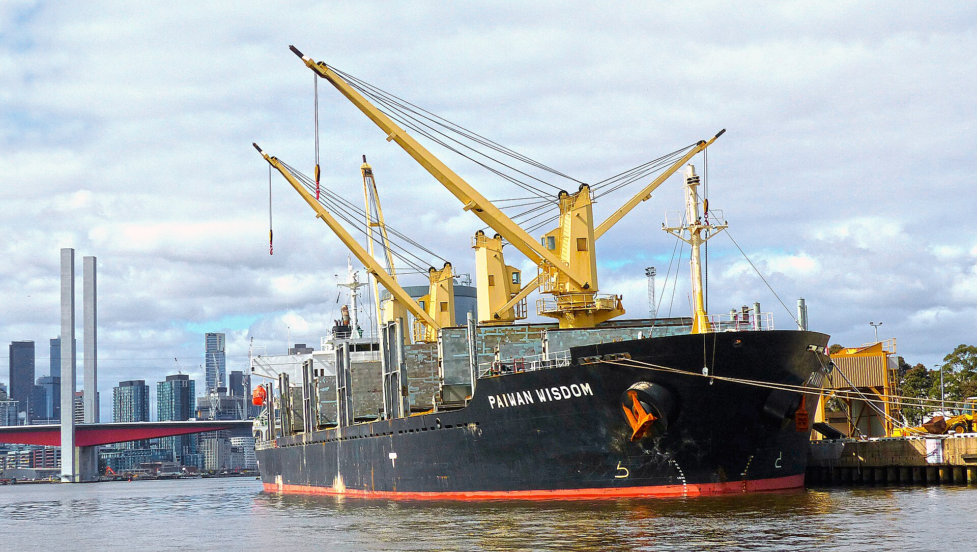 A black bulk carrier named Paiwan Wisdom with yellow cranes tied up at a port, with a city skyline and bridge behind