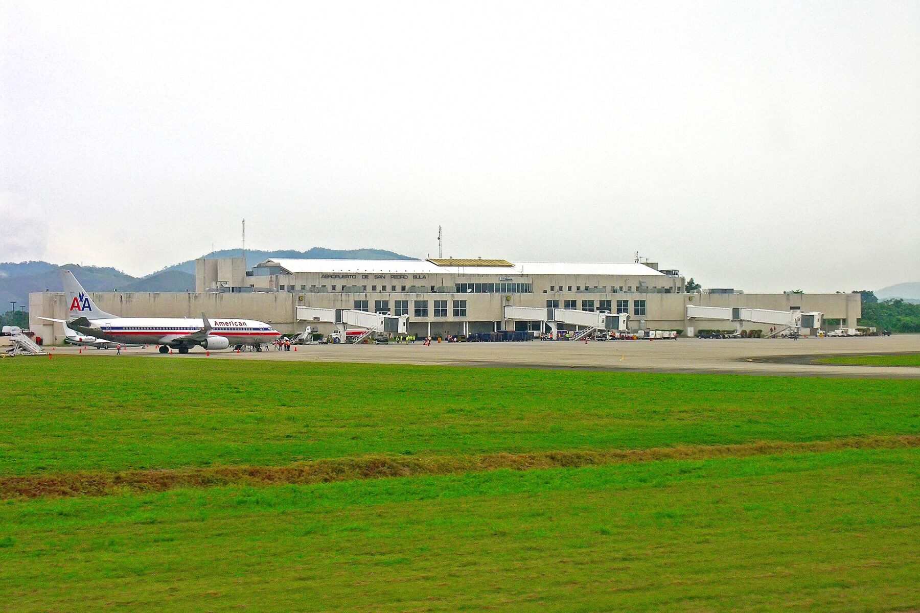 Terminal building of San Pedro Sula airport behind a grass strip, with an American Airlines jet parked at a gate on the left