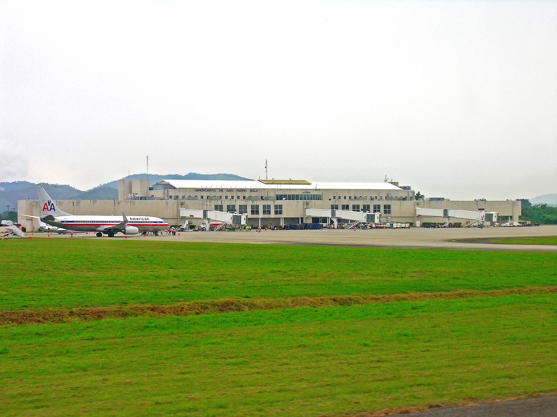 Terminal building of San Pedro Sula airport behind a grass strip, with an American Airlines jet parked at a gate on the left