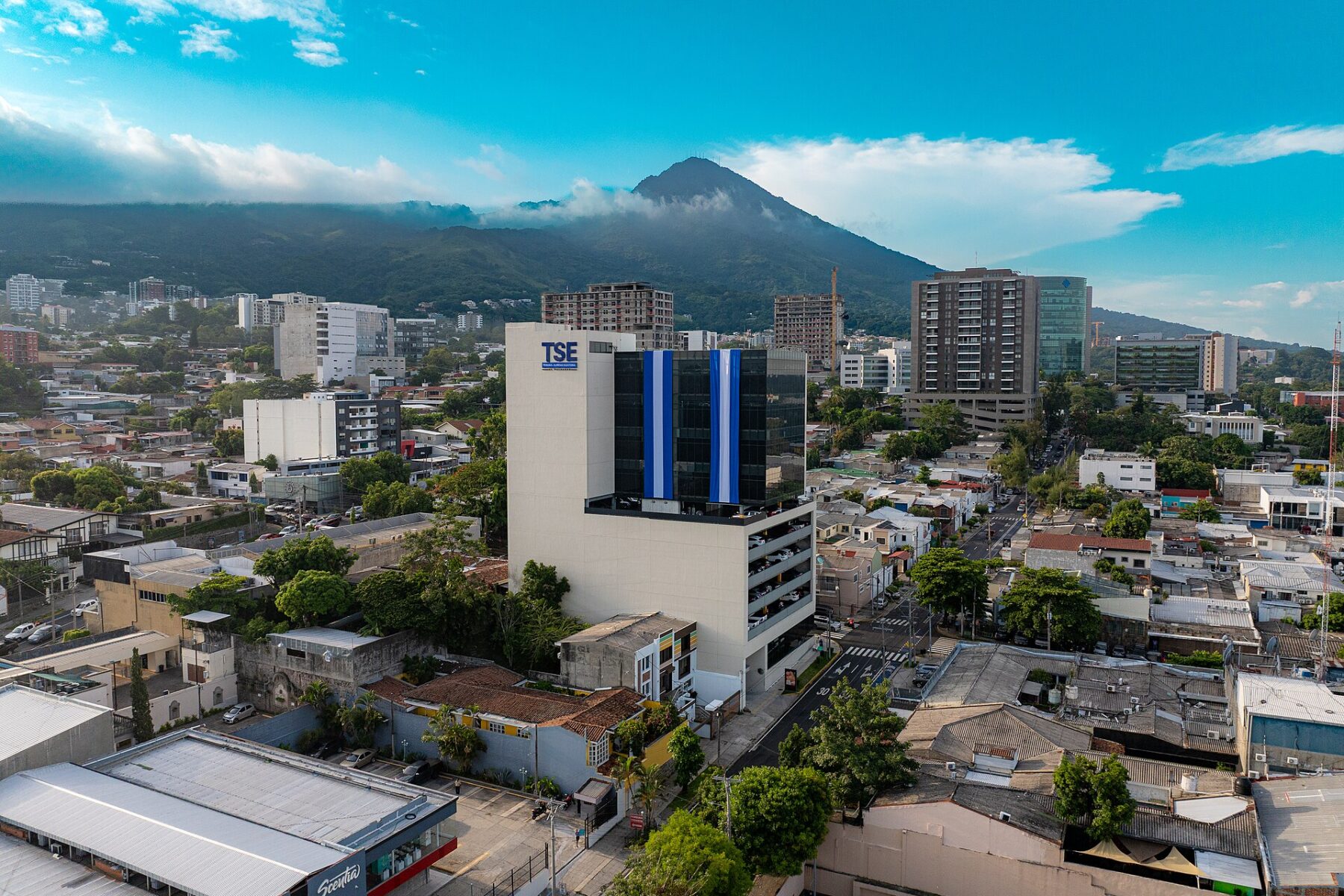 Aerial view of the Supreme Electoral Tribunal building in San Salvador with a blue-and-white striped facade, city streets and a volcano behind