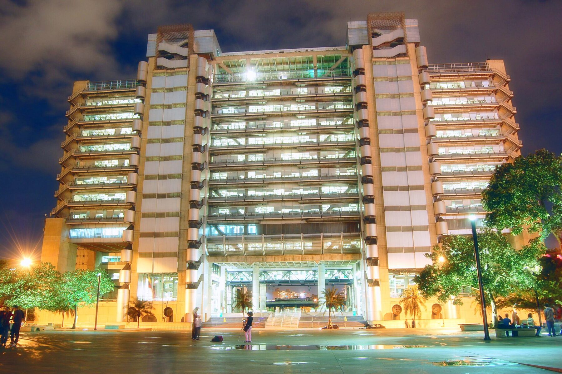 The EPM headquarters tower in Medellín, a floodlit office block with lit floors, seen at night from the plaza with people and trees in front