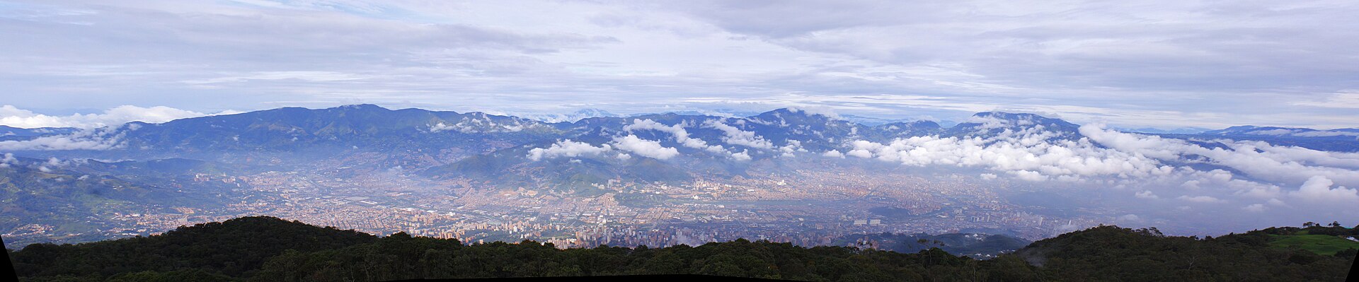 Wide panorama of the Aburrá Valley with the city spread across the valley floor below blue mountains and cloud