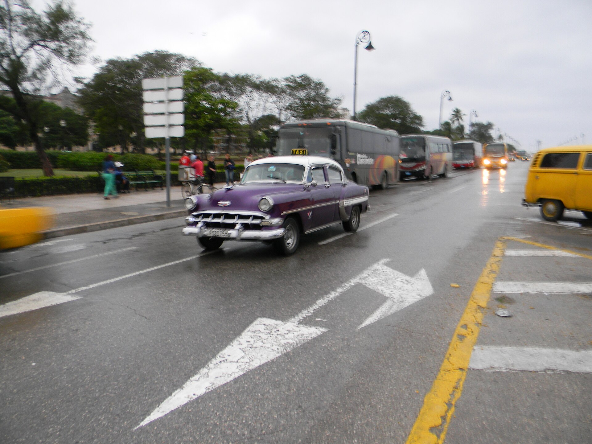 A purple and white 1950s American sedan with a taxi sign on its roof driving on a wet road, with buses behind it and a yellow van to its right