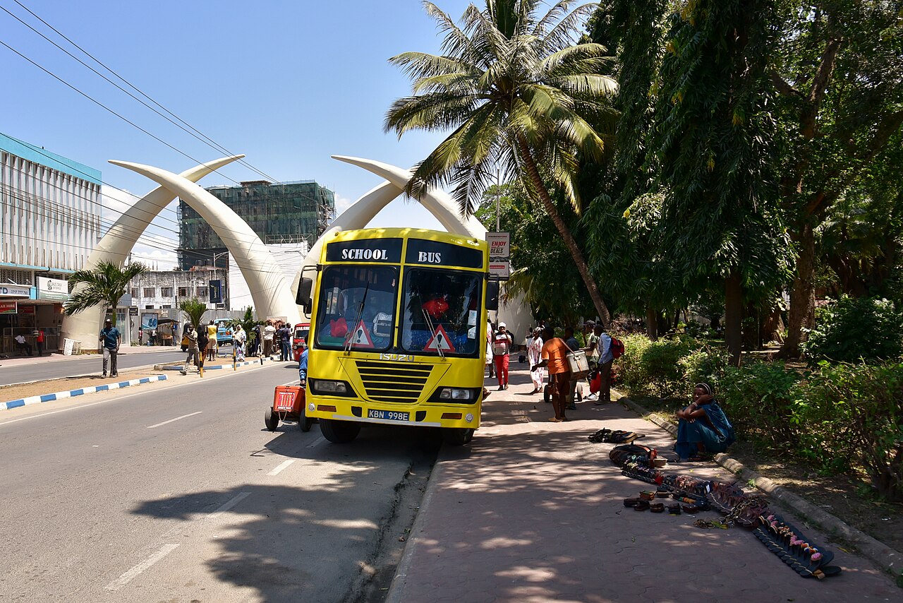 A yellow bus and pedestrians on Moi Avenue in Mombasa in front of the white tusk arches, with palm trees on the right.