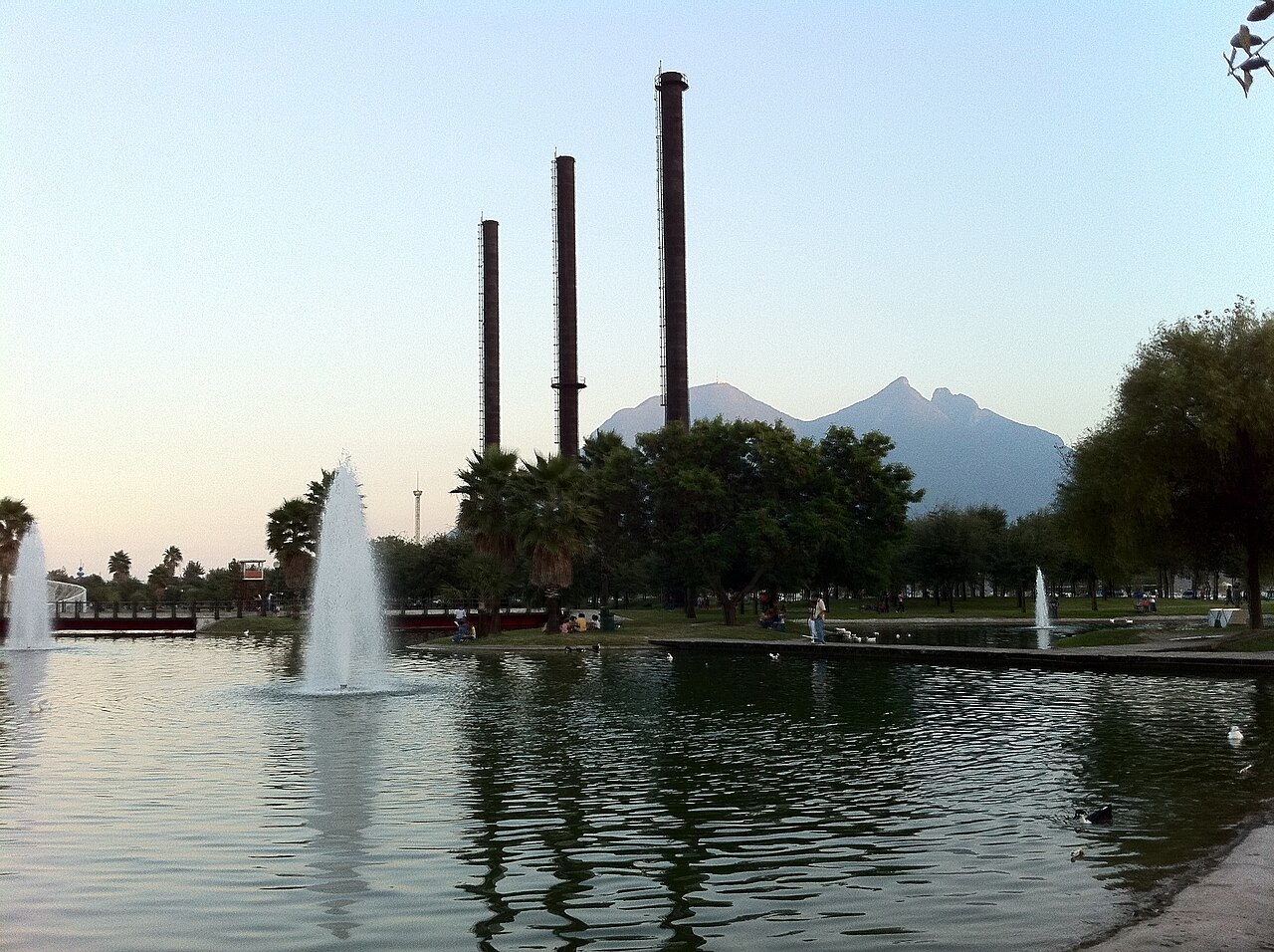A lake with fountains in Fundidora Park, three old steel-mill chimneys and the Cerro de la Silla mountain at dusk.