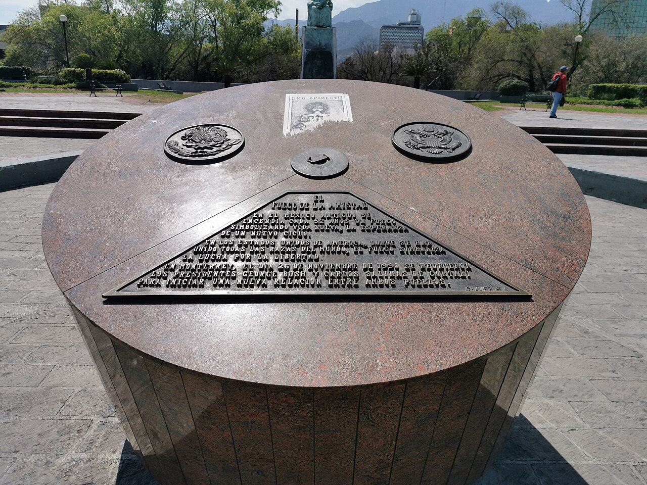 A bronze and granite monument on the Macroplaza in Monterrey with Mexican and US seals and a plaque, with mountains behind