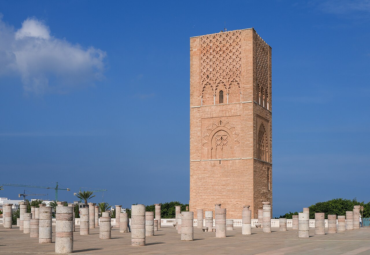 Rows of stone columns of an unfinished mosque in front of the Hassan Tower under a blue sky in Rabat, with cranes in the distance