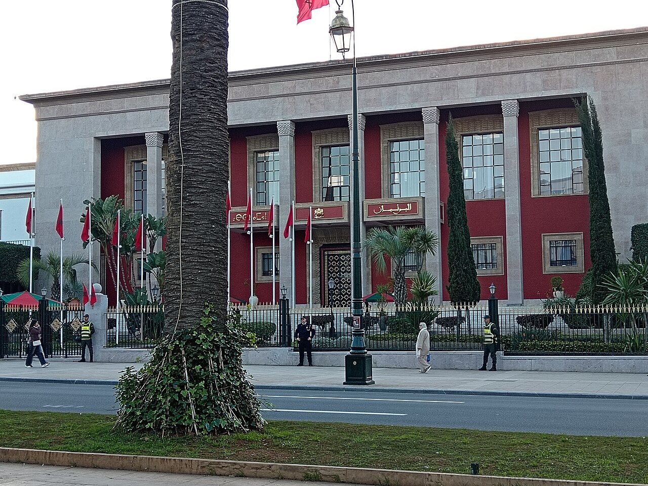 Red and stone facade of the Parliament building in Rabat, Morocco, with Moroccan flags, guards and a passer-by in front