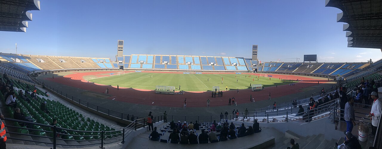 Wide view inside the Fez Sports Complex during a match, with blue and green seats and a red athletics track around the pitch