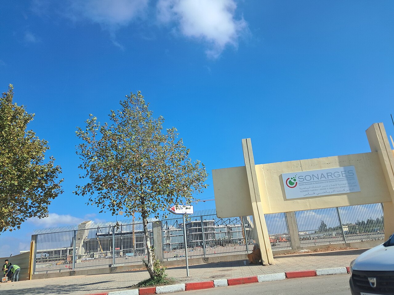 Fenced entrance of the Fez Sports Complex with a Sonarges sign, and buildings under construction behind it beneath a blue sky