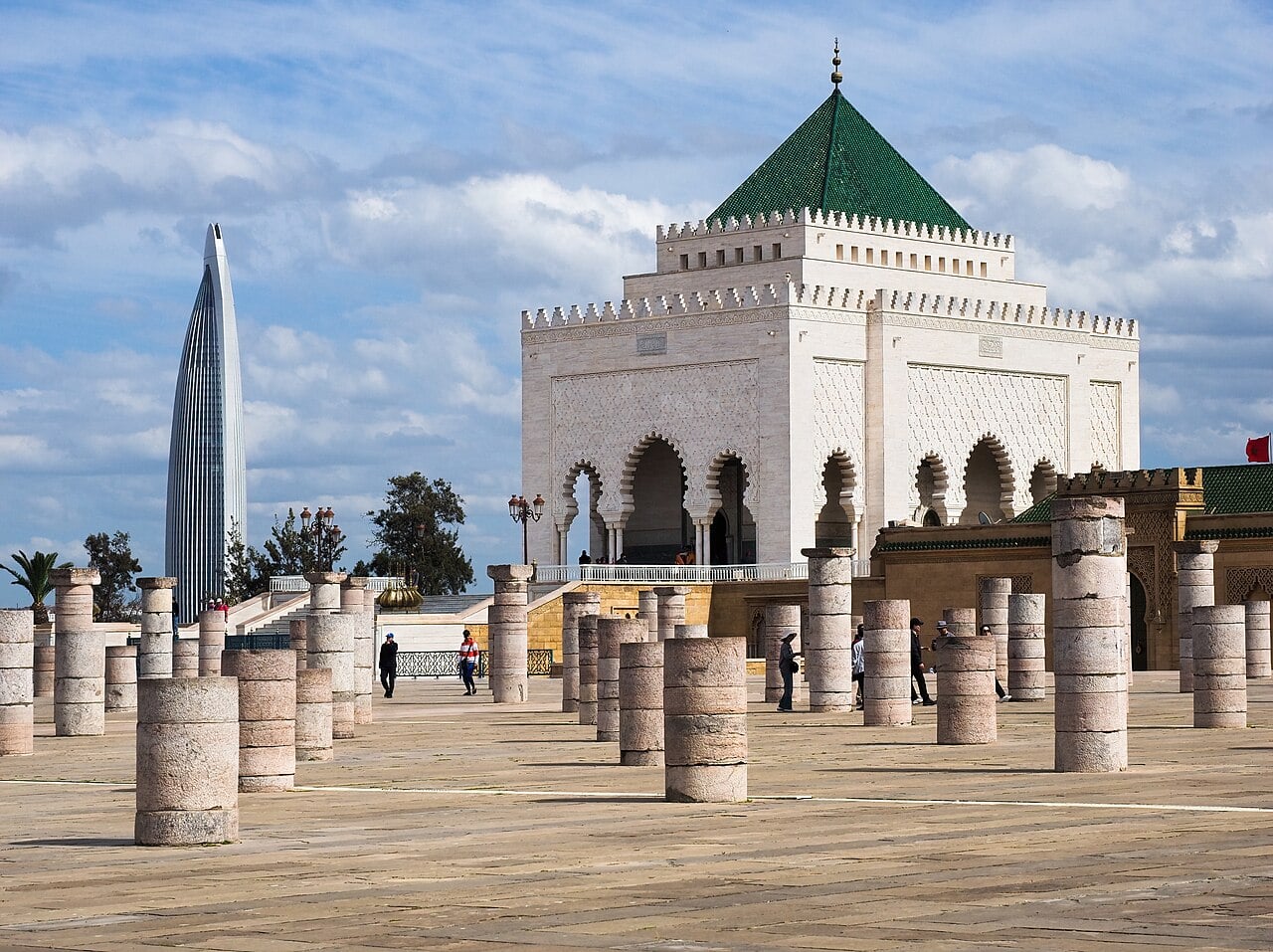 The white marble Mausoleum of Mohammed V with its green-tiled pyramid roof in Rabat, rows of stone columns in the foreground and the glass Mohammed VI Tower in the background